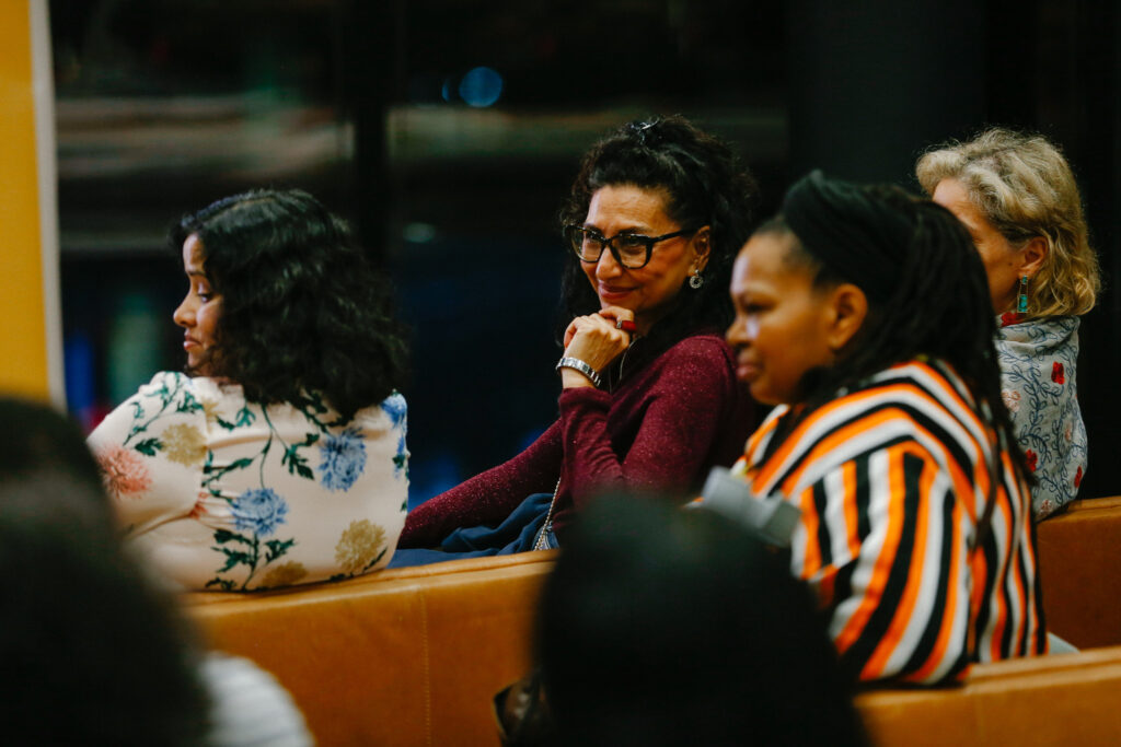 An audience image featuring 4 women in colourful clothing. 1 woman faces the camera with her hand under her chin, smiling